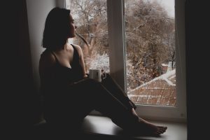 A woman looks forlorn out a window while holding a mug 