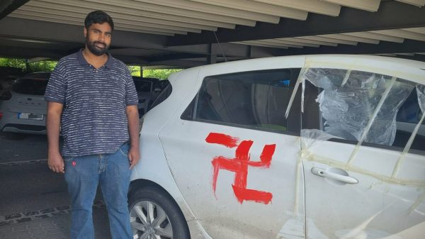 German politician standing with vandalized vehicle that has a swastika on it