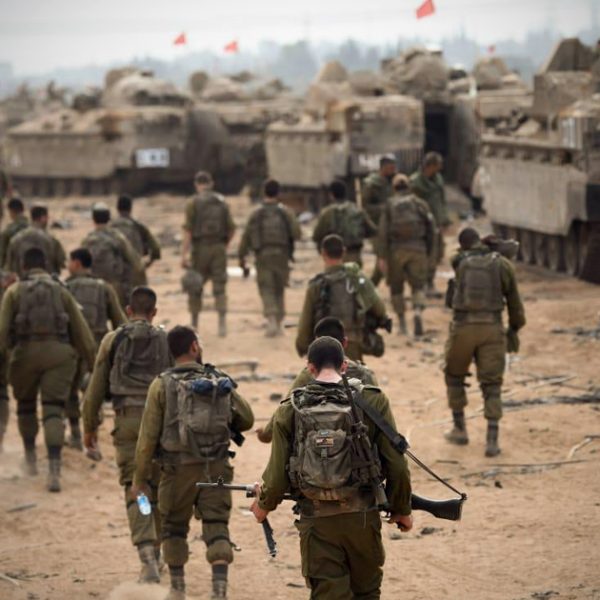 IDF soldiers walk near a group of armored vehicles on a desert ground in harsh sunlight